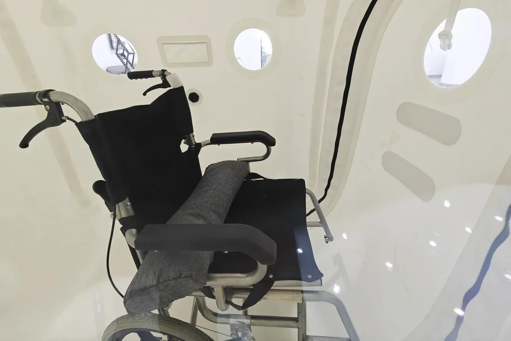 Yuvarlak lombozlu beyaz “Dominium Wellness” DW140MH model hiperbarik kabinin içinde siyah bir tekerlekli sandalye. Sahne klinik ve işlevsel bir atmosfer yansıtıyor.