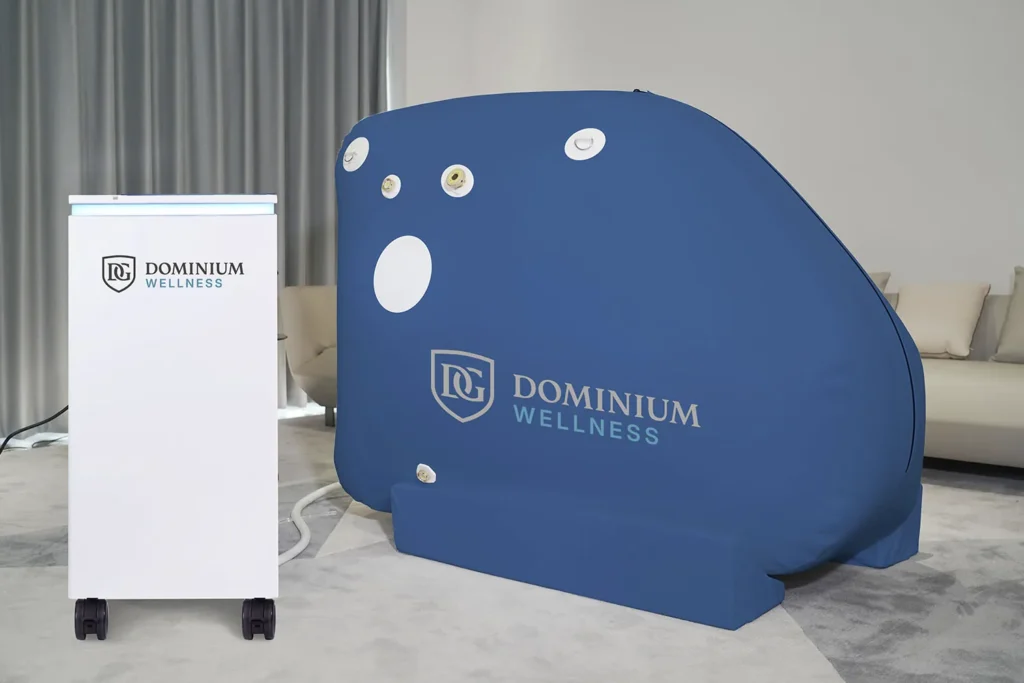 Yanında beyaz bir cihaz bulunan “Dominium Wellness” DW160MS model mavi hiperbarik kabin. Ortam perdeli ve kanepeli modern bir oda.