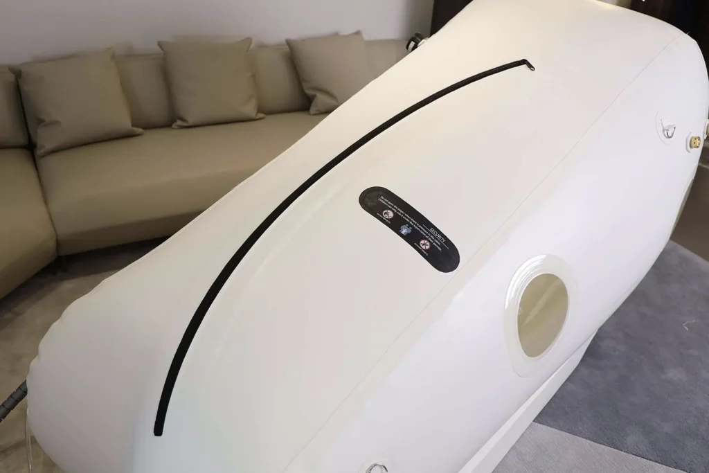 Küçük yuvarlak pencereli ve kontrol panelli “Dominium Wellness” DW220MLS model beyaz hiperbarik kabin; bej köşe koltuk ve minderlerin bulunduğu modern bir oturma odasında.
