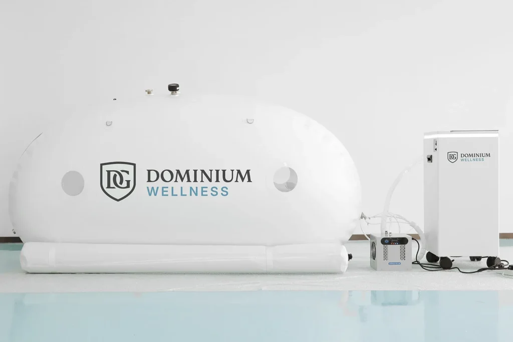 Temiz, açık mavi ve beyaz zemin üzerinde medikal ekipmana bağlı beyaz “Dominium Wellness” DW240MDO model hiperbarik kabin; klinik ve dingin bir ortam.