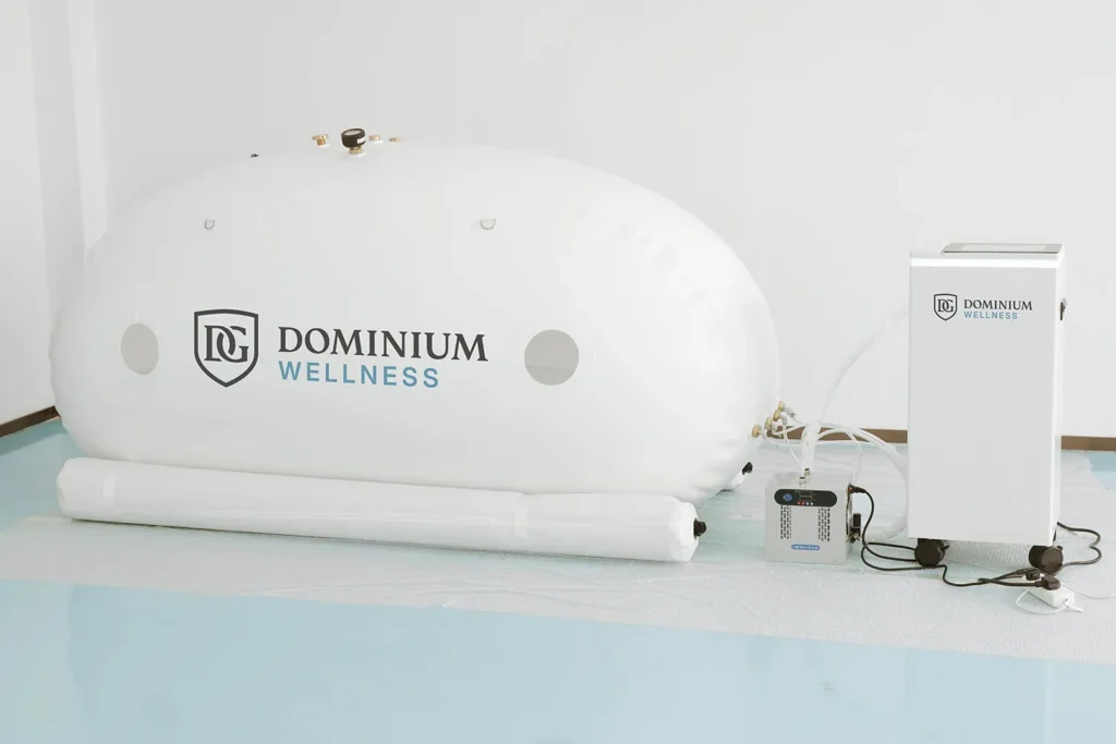 Bir kontrol ünitesinin yanında “Dominium Wellness” DW240MDO model beyaz şişirilebilir hiperbarik kabin. Ortam temiz ve minimalist; rahatlama hissi uyandırıyor.