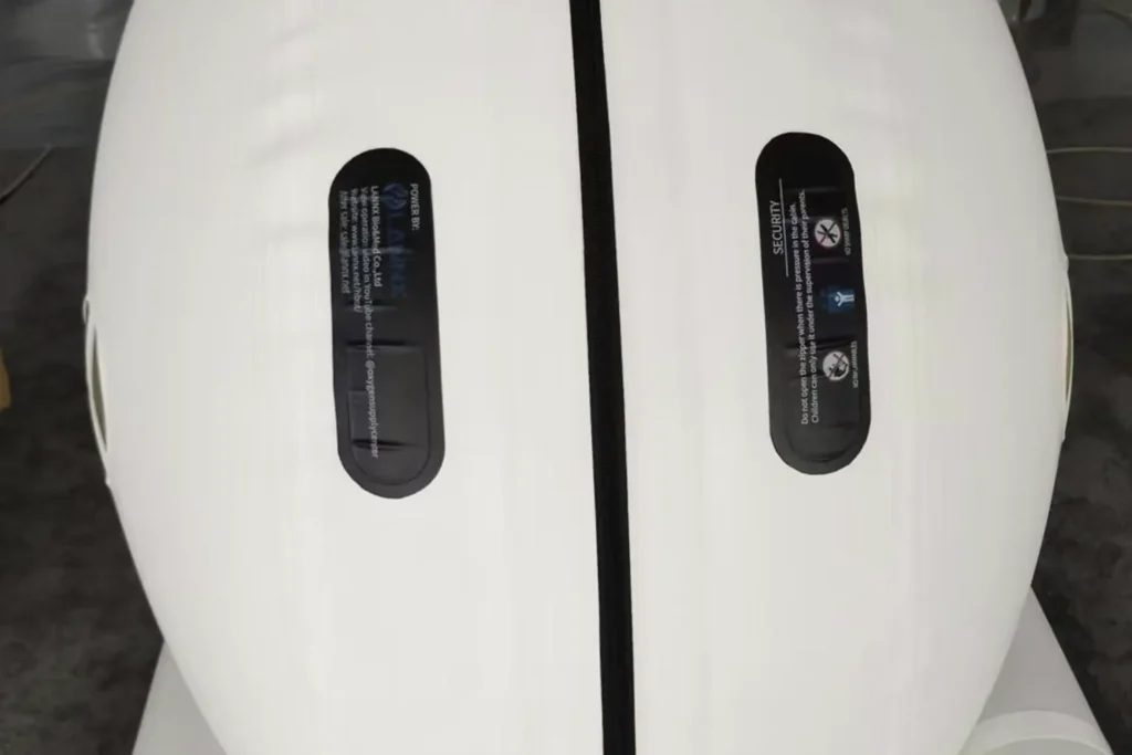 Siyah dikey şeritli beyaz “Dominium Wellness” DW240MDO model hiperbarik kabinin yakın çekimi. Yazı ve simgeler içeren iki siyah panel şık, modern bir görünüm veriyor.